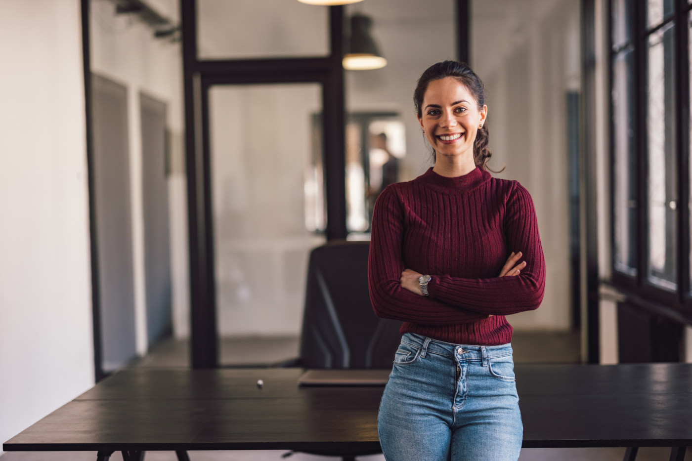 Frau lächelt in die Kamera, steht vor dem Tisch und hat die Hände verschränkt