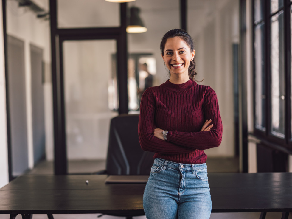 Frau lächelt in die Kamera, steht vor dem Tisch und hat die Hände verschränkt